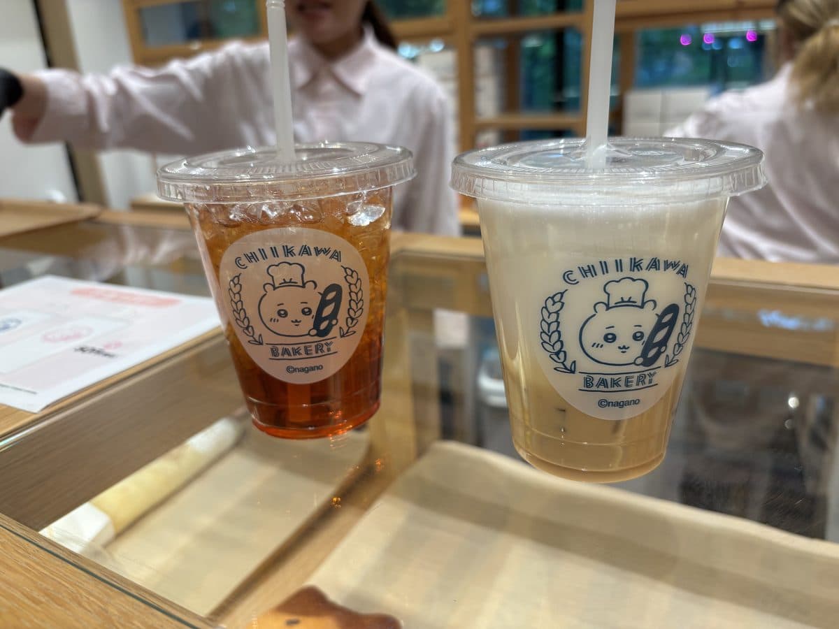 Two Chiikawa Bakery drinks — iced tea and iced café latte, held up at the ordering counter showing the branded cups and character coasters