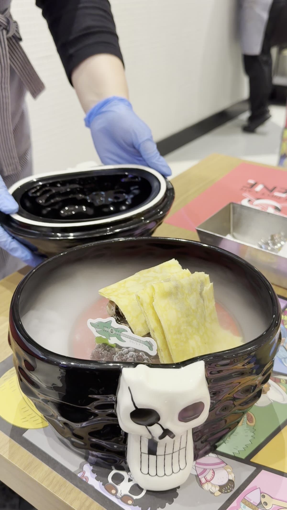 Skull bowl ramen being served at One Piece Cafe GENE, staff lifting the Trafalgar Law Jolly Roger lid to reveal salt ramen inside