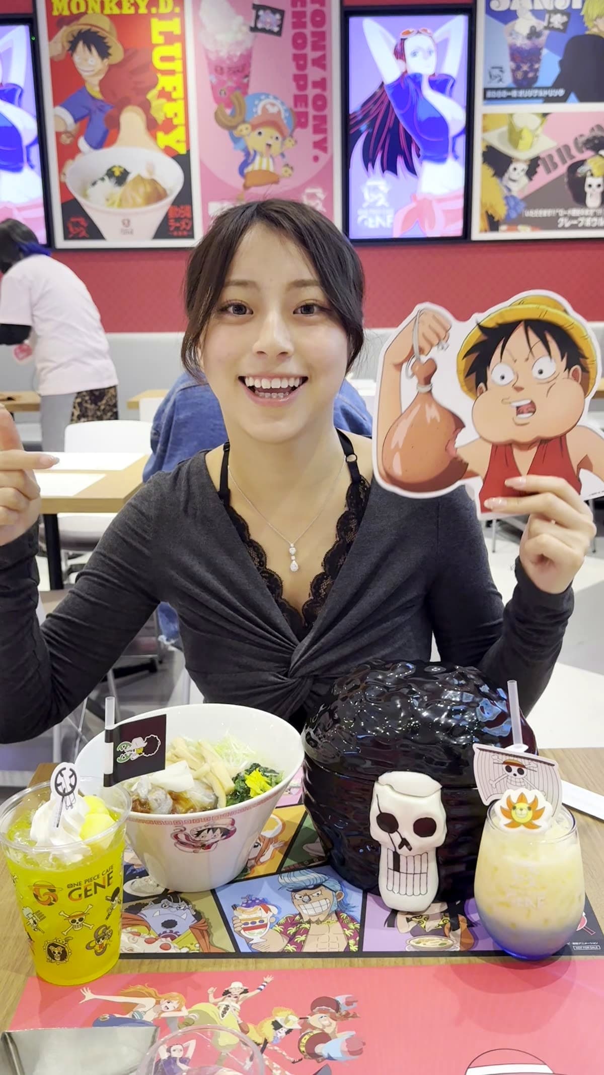Moe from Japan Pop Now with full table spread at One Piece Cafe GENE Shibuya, skull bowl ramen, crepe bowl, two character drinks, Luffy standee, and Straw Hat Crew placemat