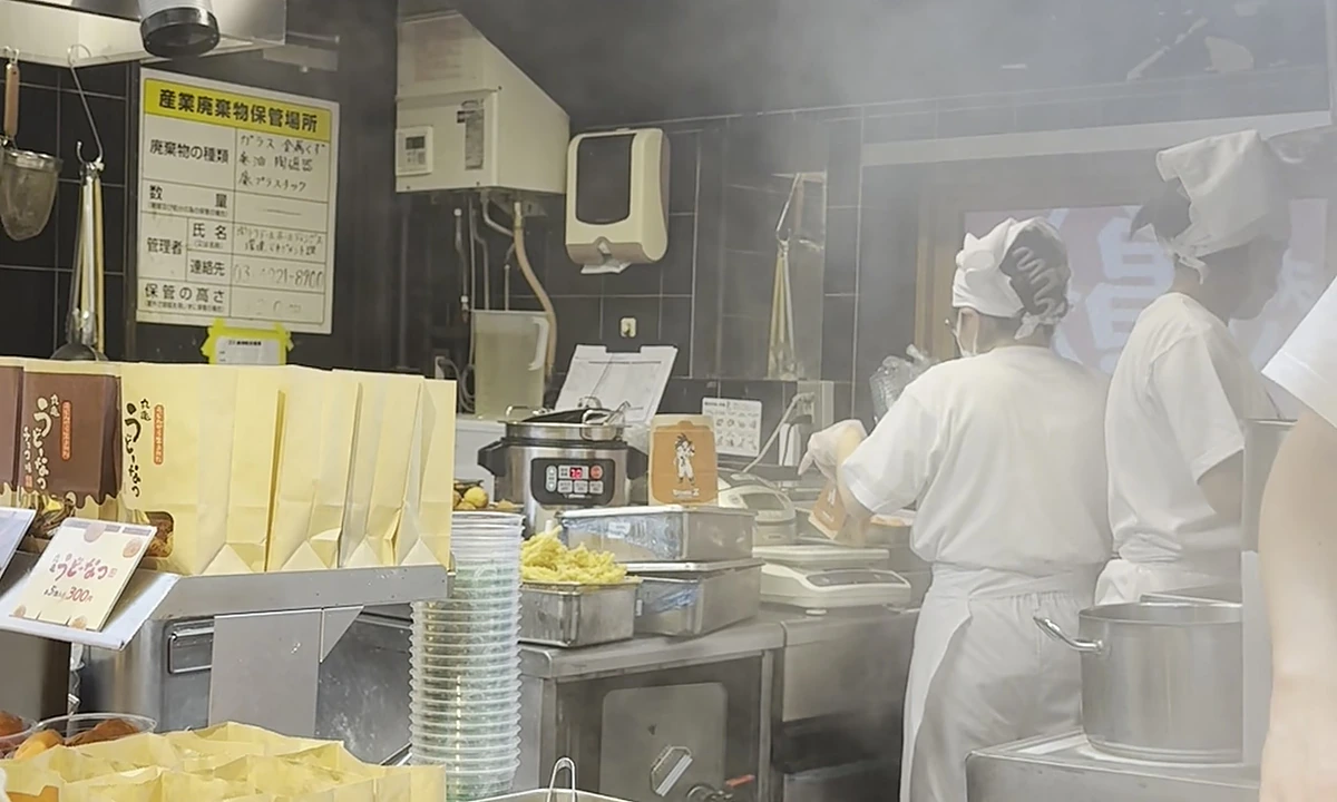 Marugame kitchen interior with Senzu tempura takeout bags lined up in foreground tray, white-uniformed staff working steamer/fryer equipment in background, ventilation hoods visible