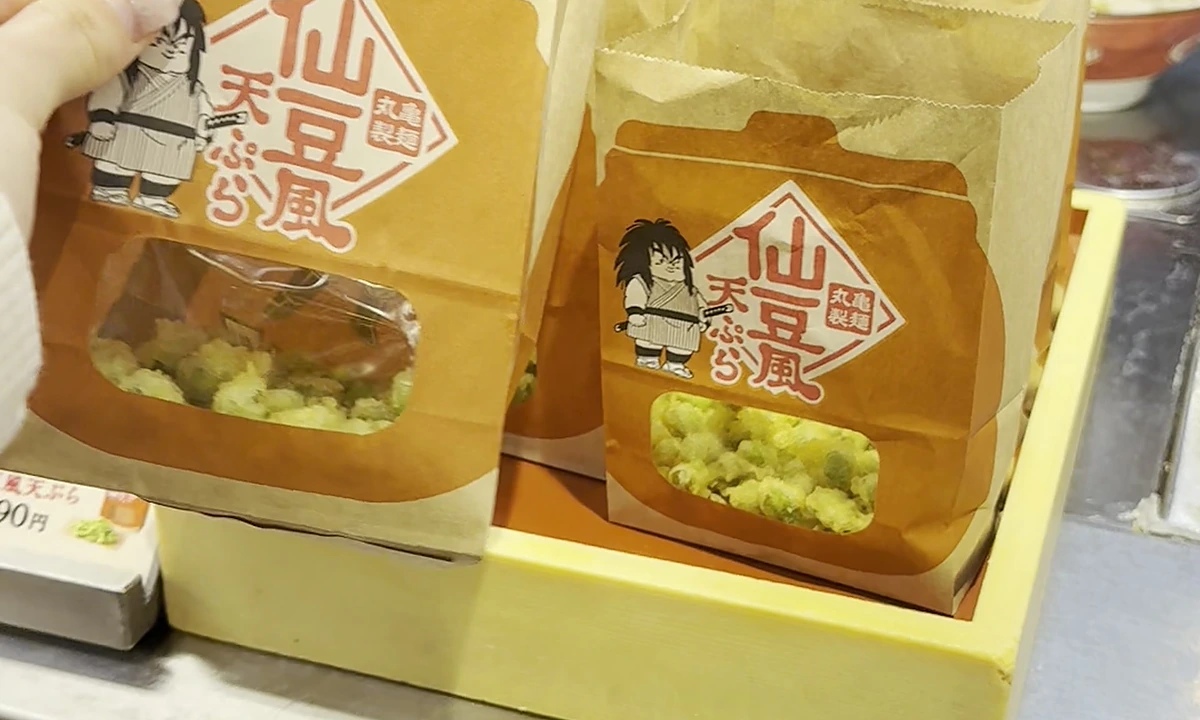 Two Senzu-style tempura takeout bags with character art (Yamcha-style and Trunks-style) on the front, edamame visible through the cellophane window, Marugame Seimen logo top-left of each bag