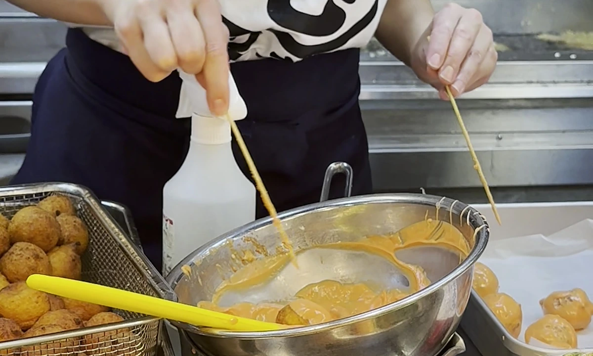 Marugame staff member in branded Dragon Ball Z collab T-shirt working a deep-fryer station, with cooked tempura and udon-uts on trays in the foreground