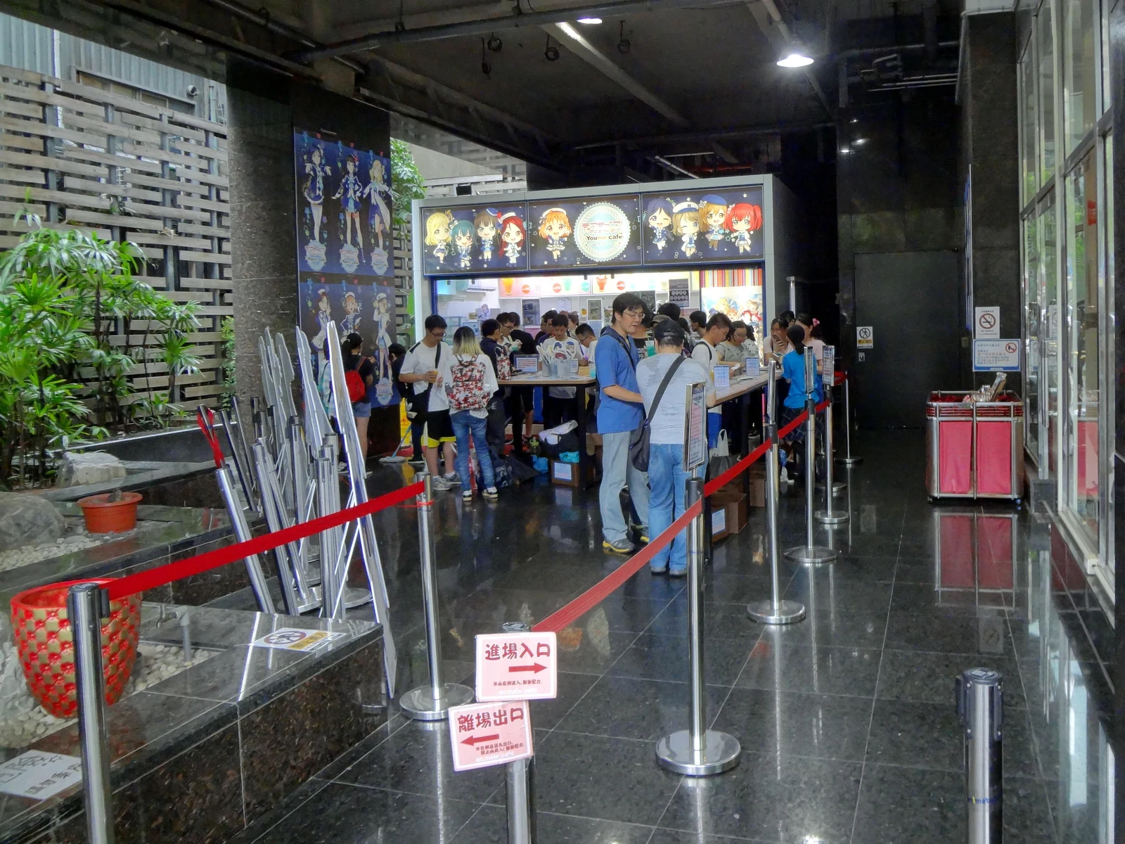 Inside an anime collaboration cafe, character standees, merch displays, and the queue rail with red rope barriers leading to the order counter