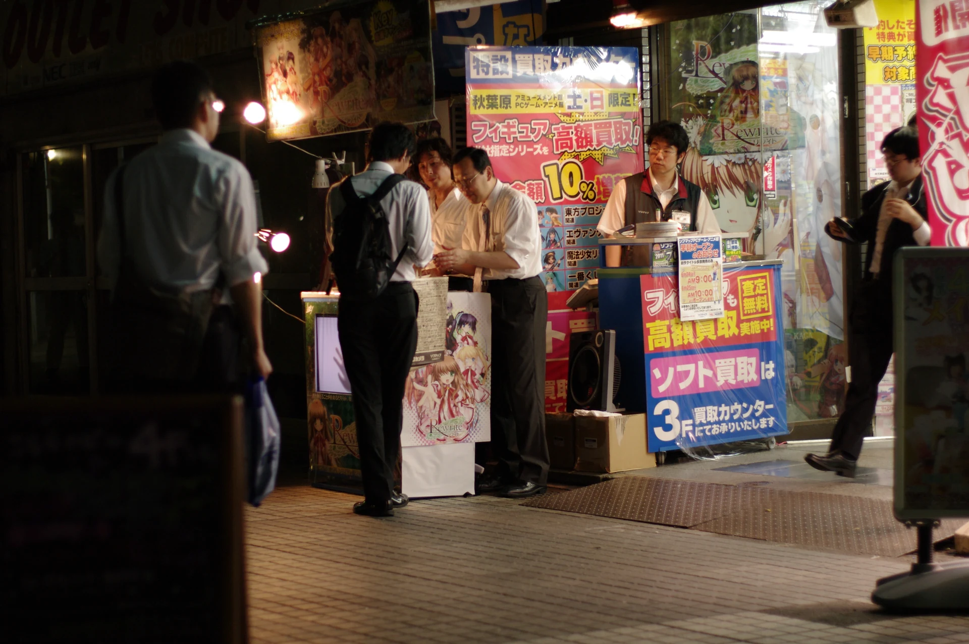 Customers gathered at a Sofmap Akihabara PC Game and Anime Museum shop counter at night, with promotional posters lining the entrance, a typical late-evening figure-hunting scene proxy buyers ask shops to ship from