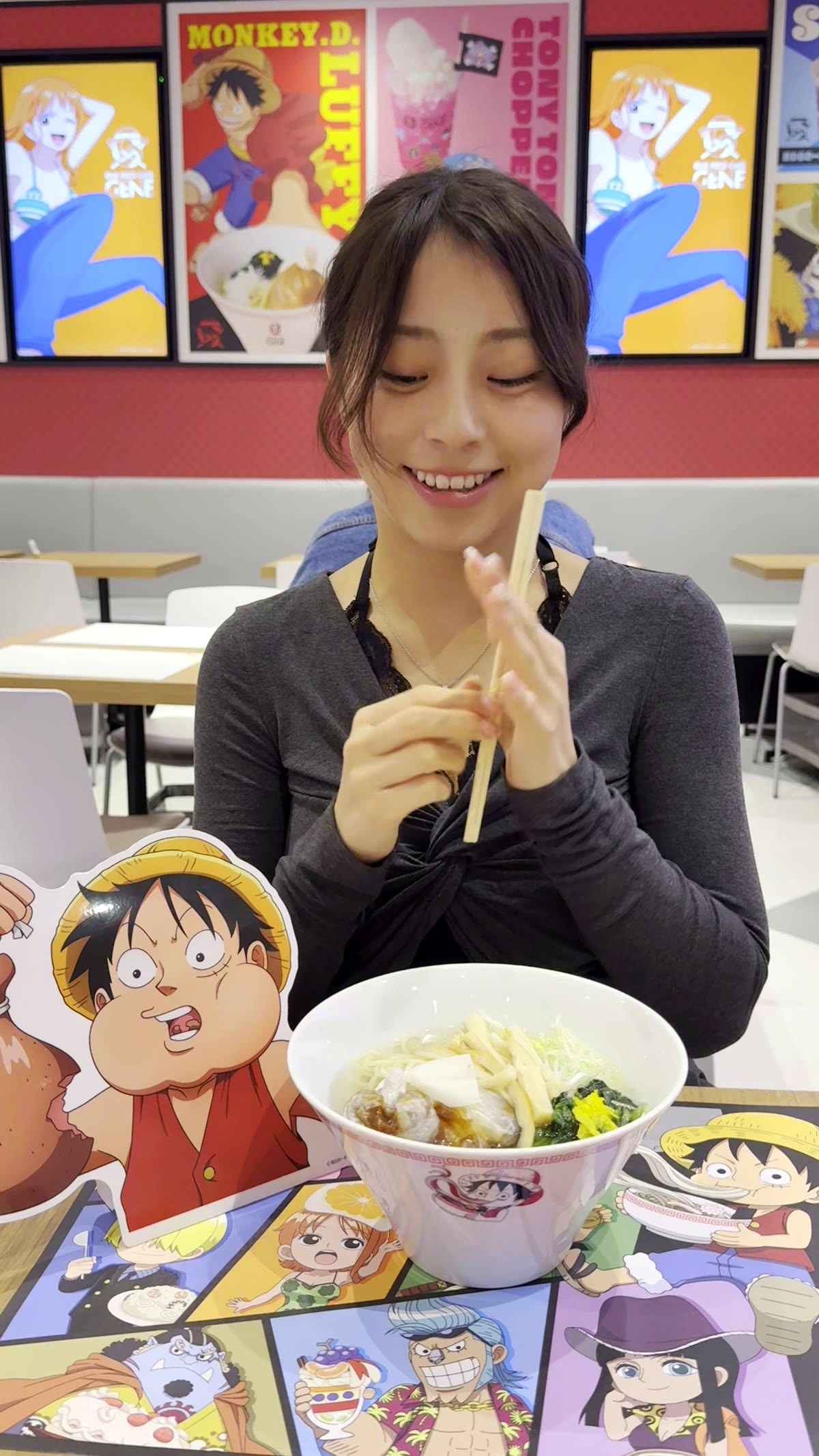 Moe from Japan Pop Now eating Straw Hat Salt Ramen with chopsticks at One Piece Cafe GENE, with Luffy character standee and colorful One Piece posters in the background