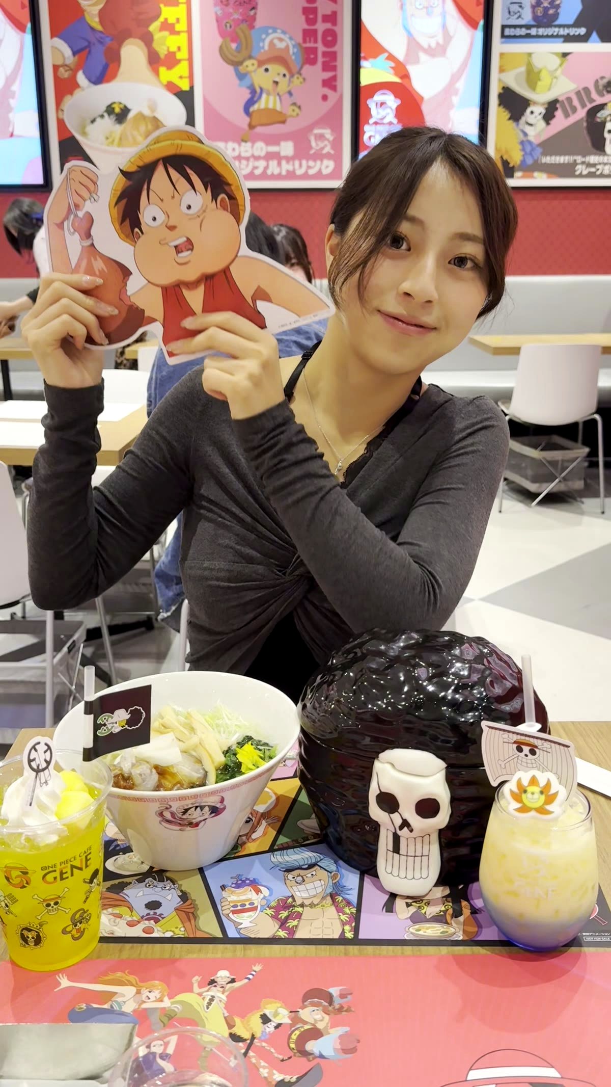 Moe from Japan Pop Now holding a Luffy cardboard standee at One Piece Cafe GENE with skull bowl ramen, character drinks, and birthday cards spread across the table