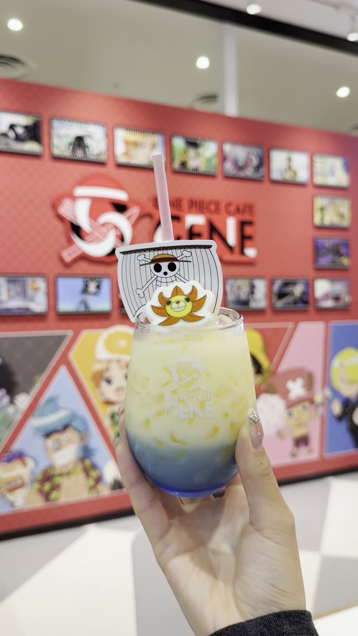 Thousand Sunny drink held up in front of the One Piece Cafe GENE red logo wall — blue and yellow layered drink with skull Jolly Roger topper